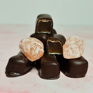 A tempting pyramid of Poppy's Chocolate Coated Turkish Delight Dark Chocolate 100g chocolates, with one piece split in half to showcase its caramel-like center, sits attractively on the soft pink surface—an ideal gift. Nearby lie two powdered sugar-coated candies evoking the traditional taste of Turkish Delight.