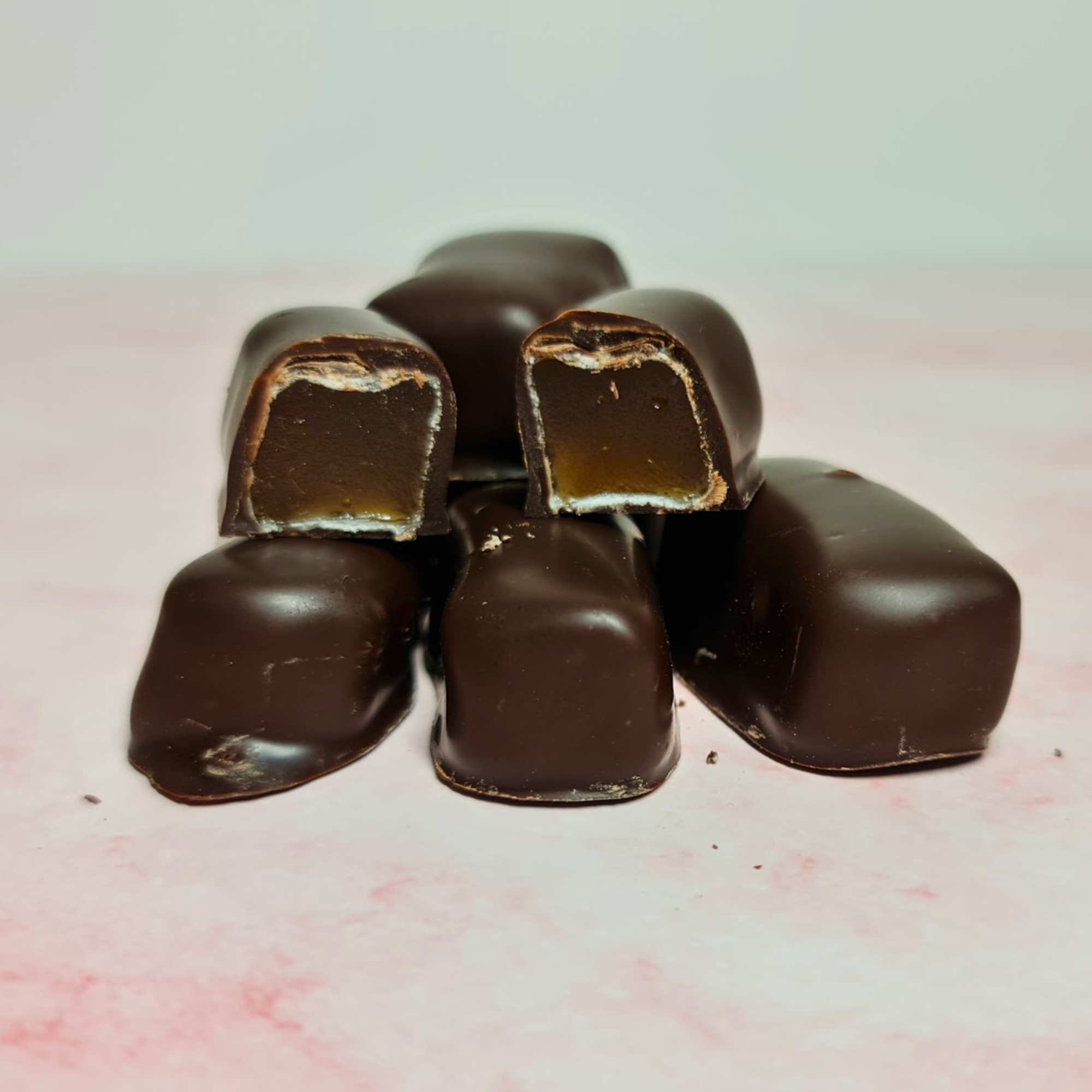 A tempting pyramid of Poppy's Chocolate Coated Turkish Delight Dark Chocolate 100g chocolates, with one piece split in half to showcase its caramel-like center, sits attractively on the soft pink surface—an ideal gift. Nearby lie two powdered sugar-coated candies evoking the traditional taste of Turkish Delight.