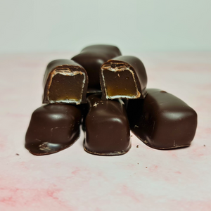 A stack of Poppy's Chocolate Coated Turkish Delight Dark Chocolate 100g is arranged on a pink surface, making it an ideal gift. One delight is cut in half, showcasing the caramel-like interior. The glossy dark chocolate coating beautifully contrasts with the smooth filling.